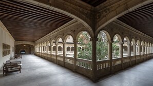Um corredor coberto espaçoso, com colunas e arcos de pedra, bancos ao longo das paredes e vista para o exterior.