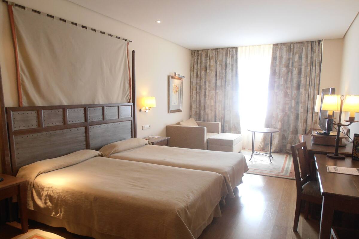 A hotel room with two beds, a desk, a chair, a small table, and a lamp.