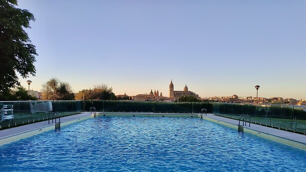 A rectangular swimming pool with clear blue water, surrounded by a glass railing. In the background, there is a cityscape with historical buildings and a clear sky.