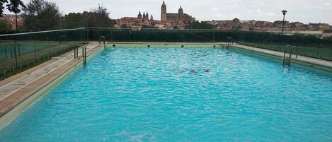 Una piscina con agua azul cristalina, rodeada por una cerca y con vista a un edificio histórico al fondo.