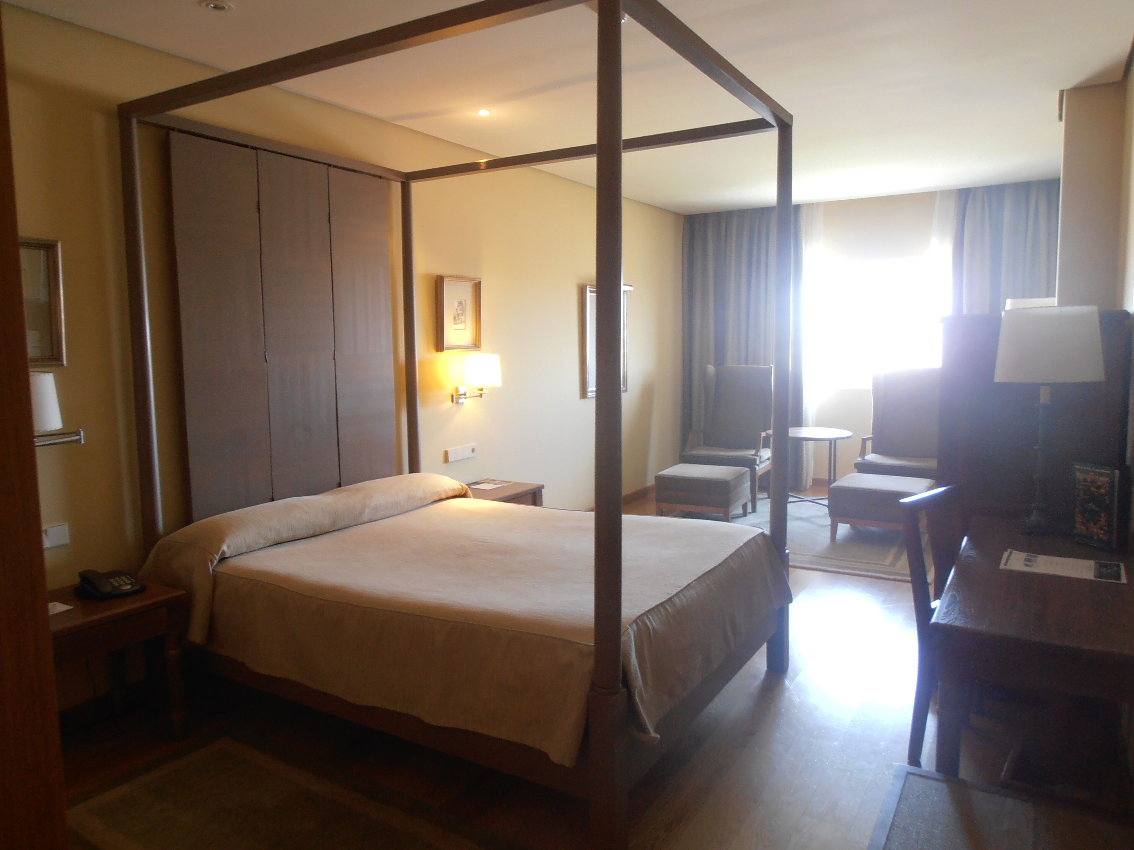 A four-poster bed in a hotel room with a desk, chair, and a small table.