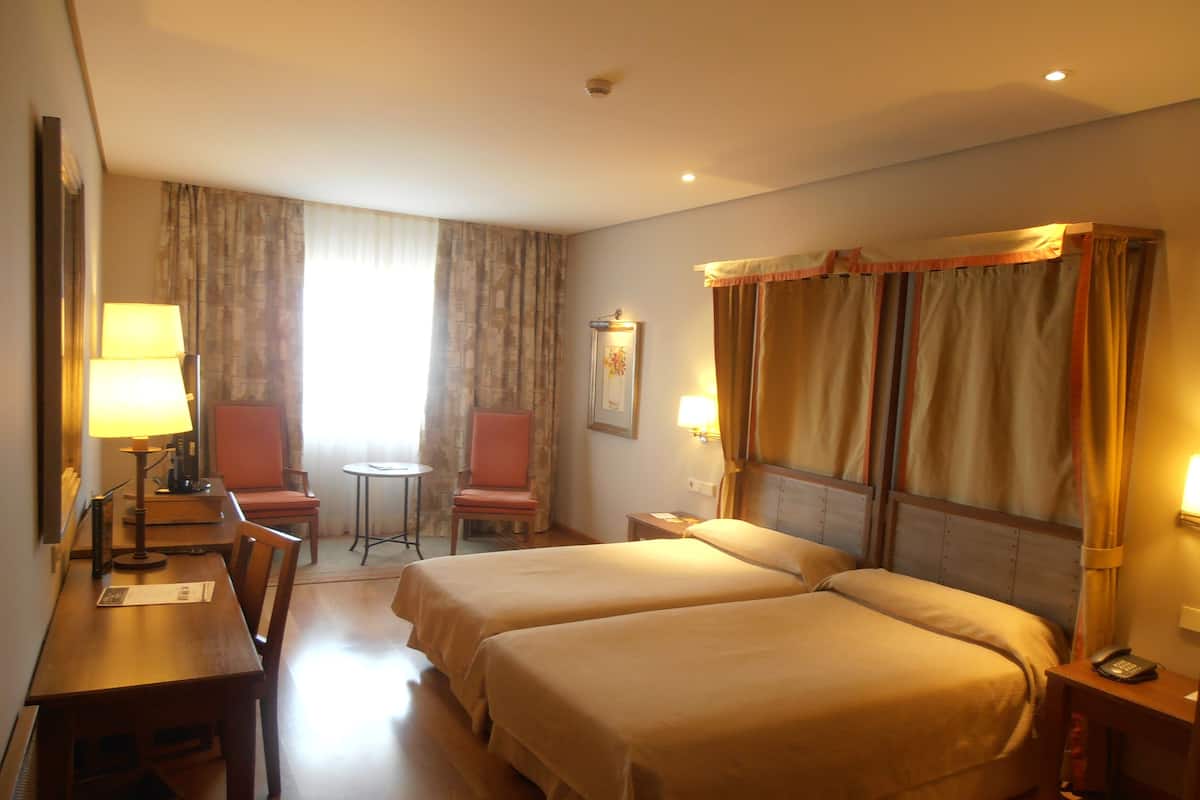 A hotel room with two beds, a desk, a chair, a small table, and a window with curtains.