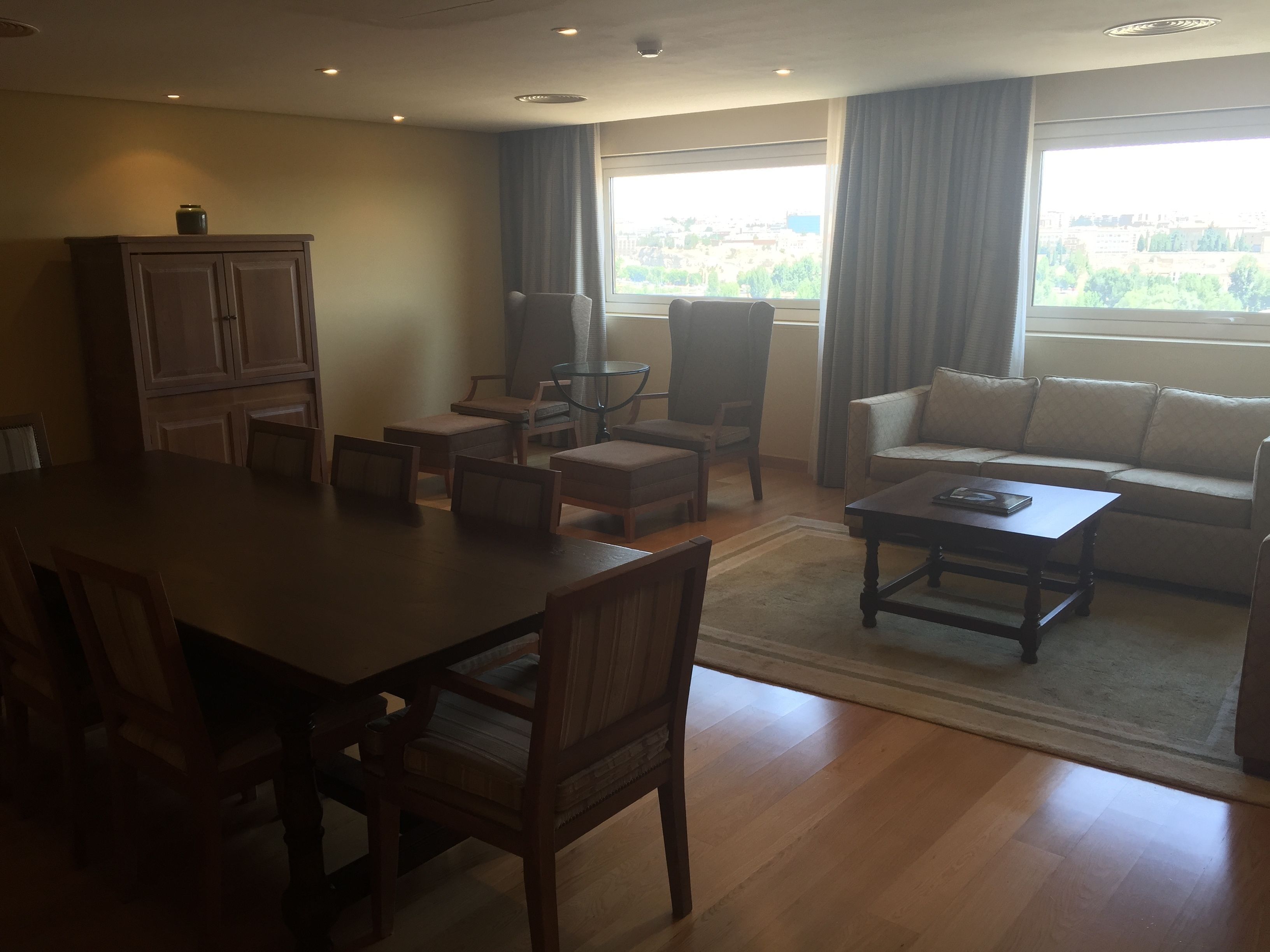 A spacious living area with a dining table, multiple chairs, a sofa, and a coffee table. There is a large window providing a view of the cityscape.