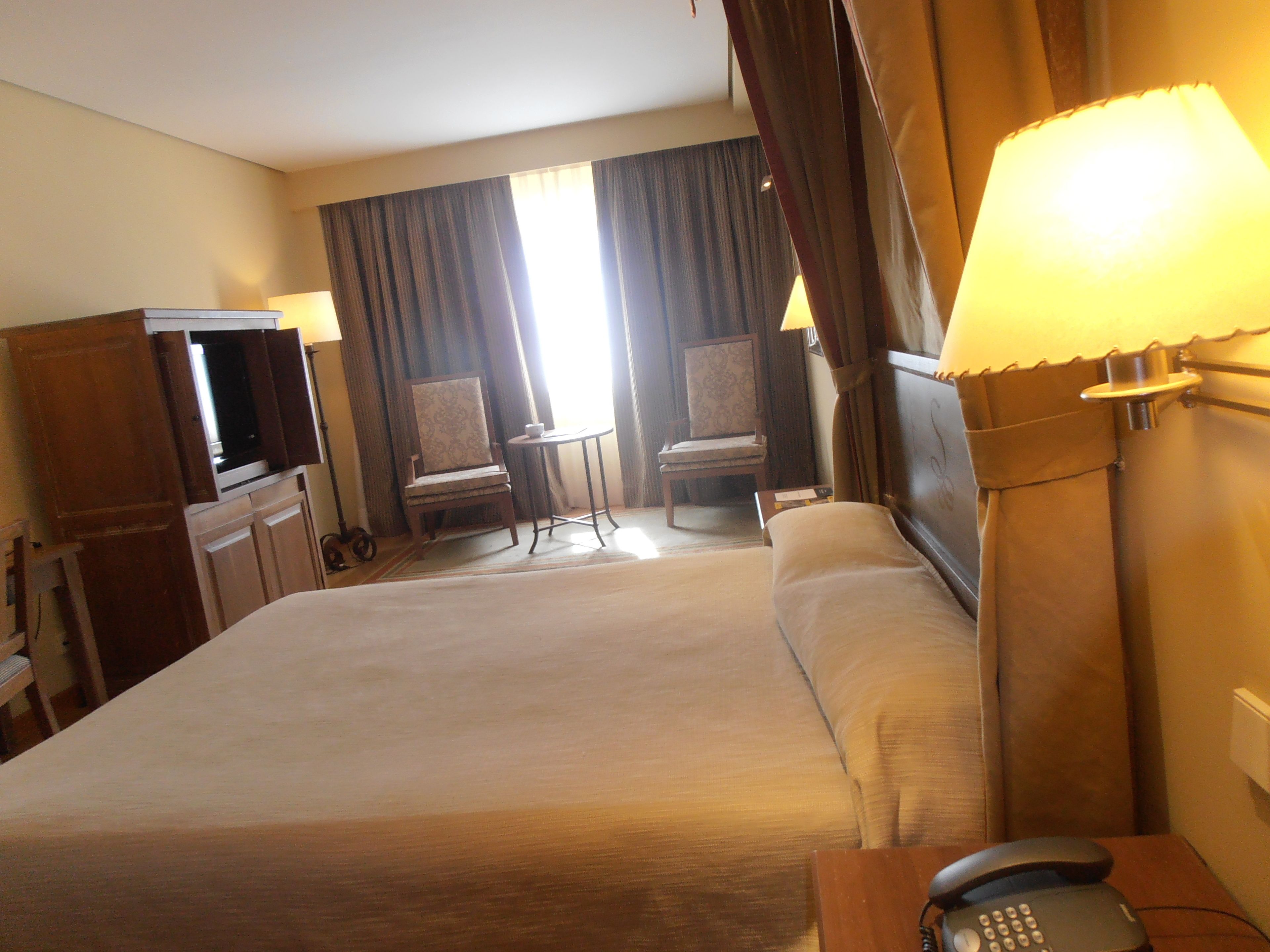 A hotel room with a large bed, a desk with a telephone, a television, two chairs, a small table, and curtains.