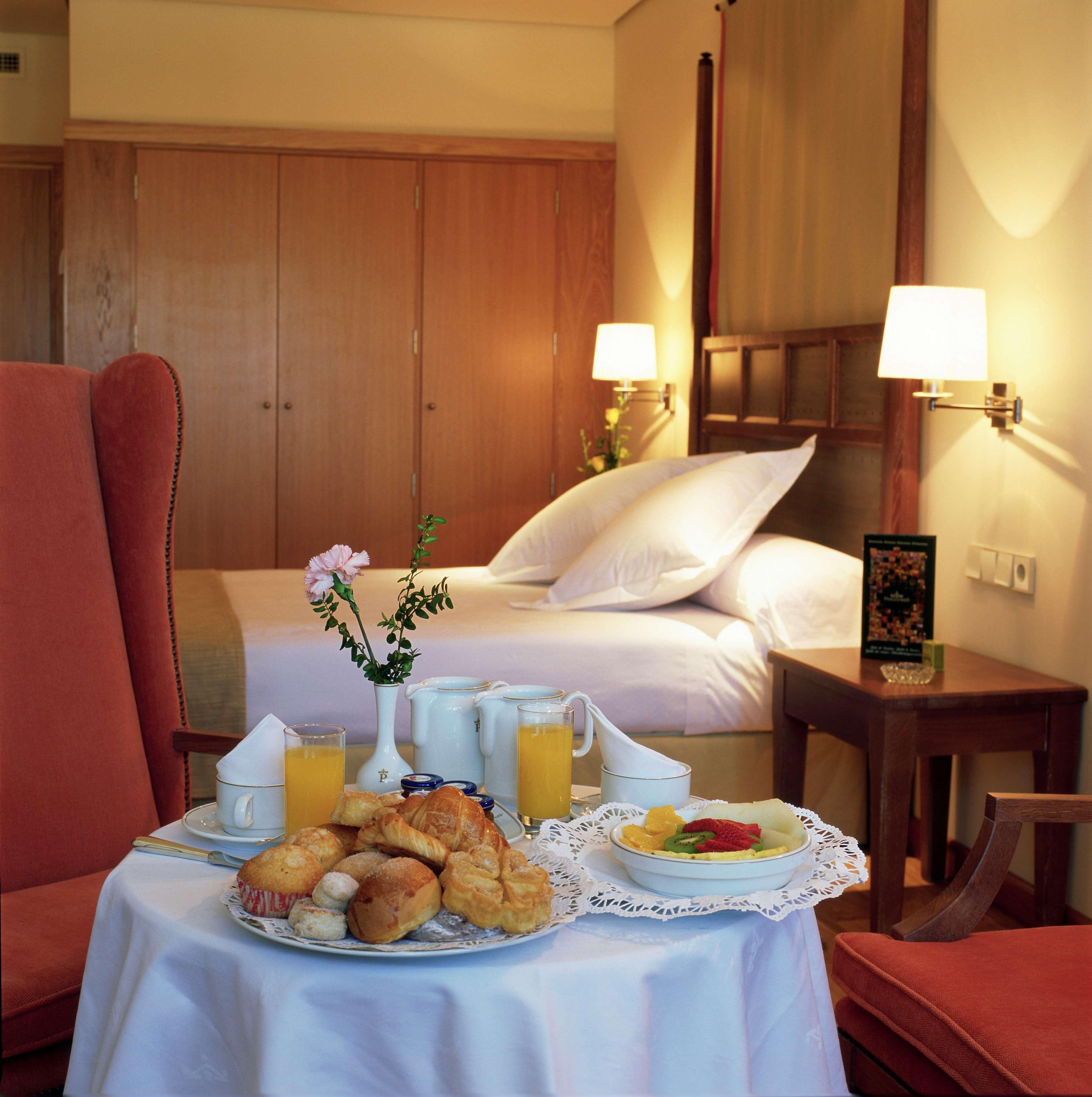 Quarto de hotel com cama, mesa de café da manhã, vaso de flores e duas luminárias.