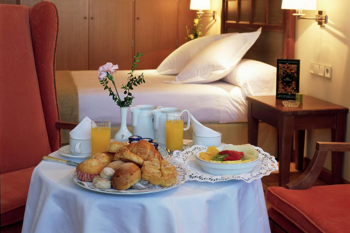 Quarto de hotel com cama, mesa de café da manhã, vaso de flores e duas luminárias.