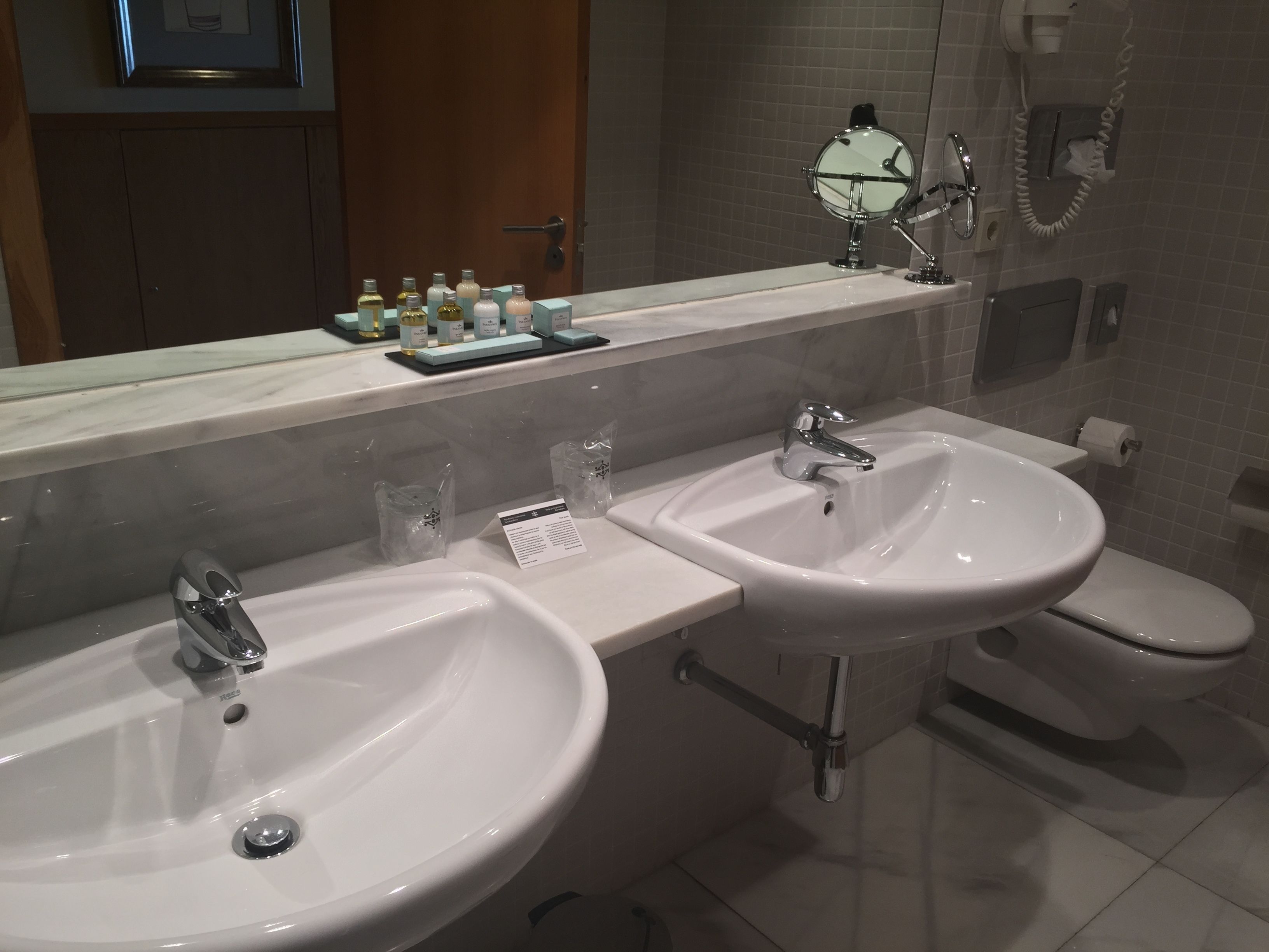 A bathroom with two sinks, a toilet, and a mirror. There are toiletries on the counter.