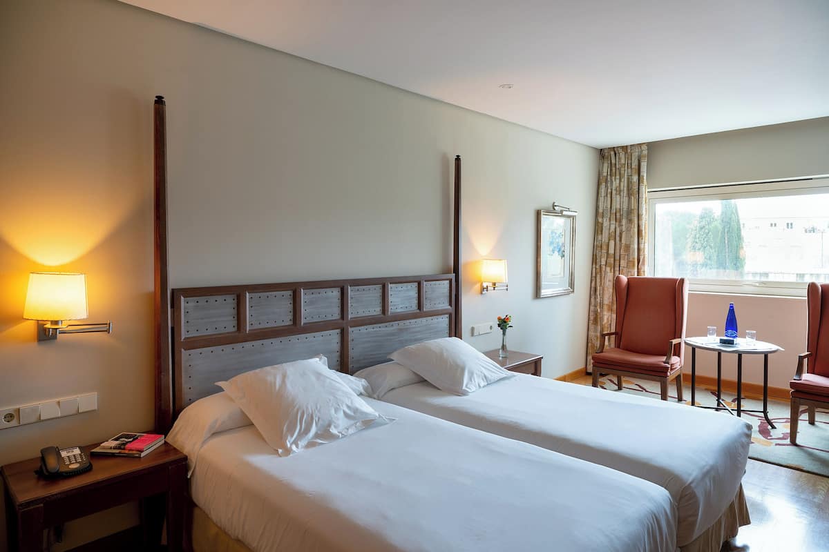 Quarto de hotel com duas camas, uma mesa de cabeceira, luminária, cadeira, mesa e janela.