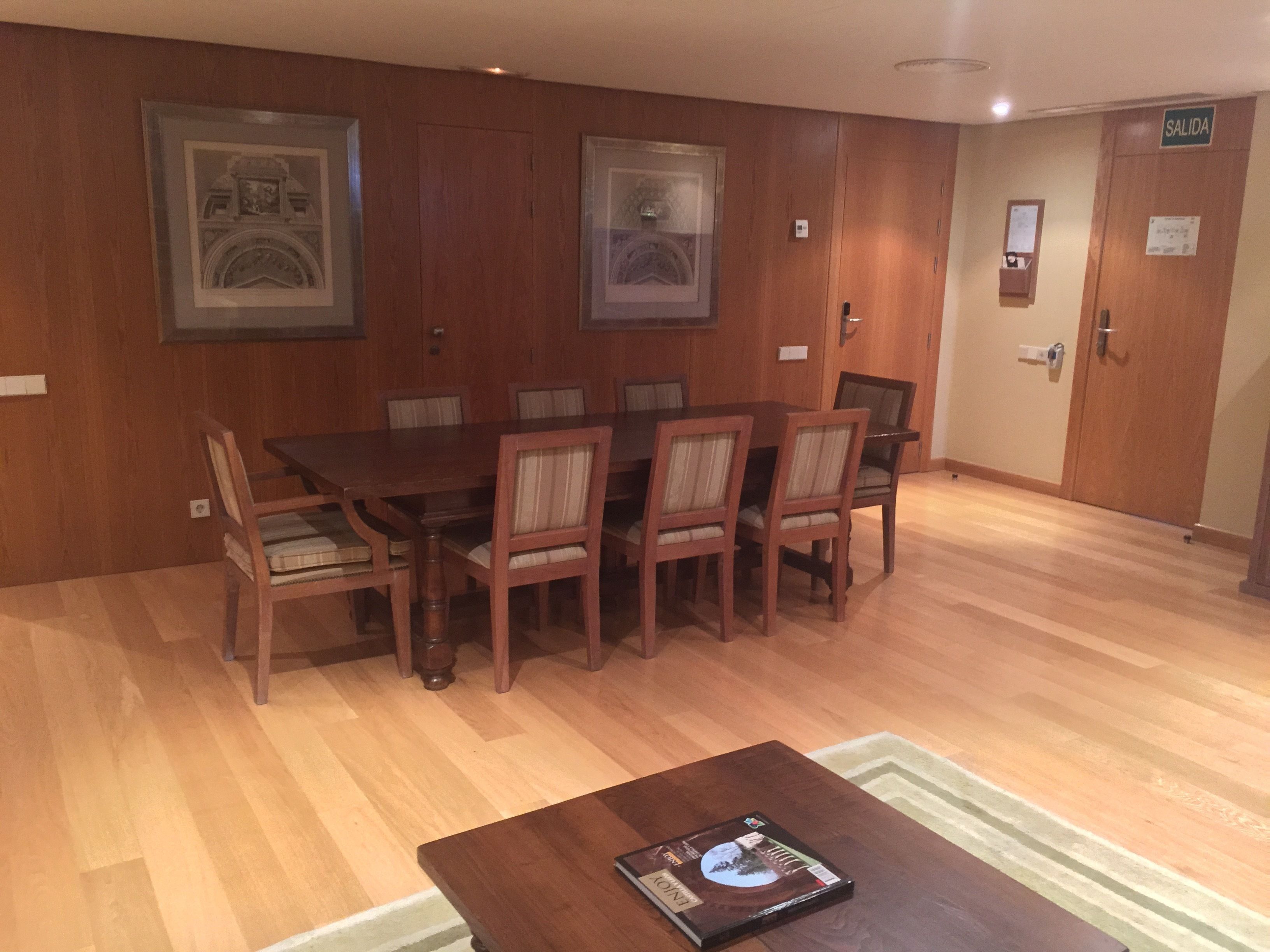 A dining area with a wooden table and chairs, framed pictures on the wall, and a door labeled "SALIDA".