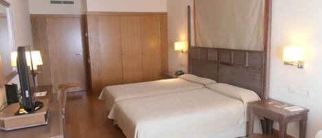 A hotel room with two beds, wooden wardrobe, bedside lamps, and a desk with a lamp.