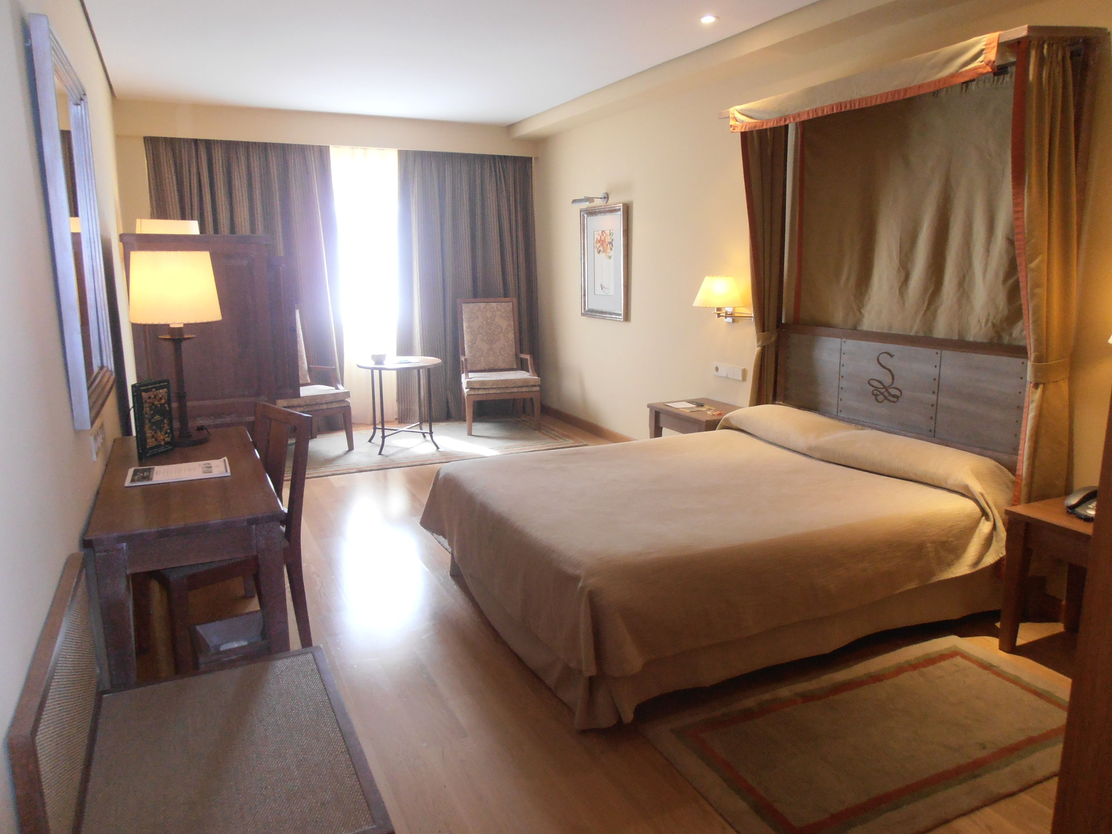 A hotel room with a large bed, a desk with a chair, a small table, and a chair. There is a window with curtains, a wall-mounted lamp, and a picture frame.