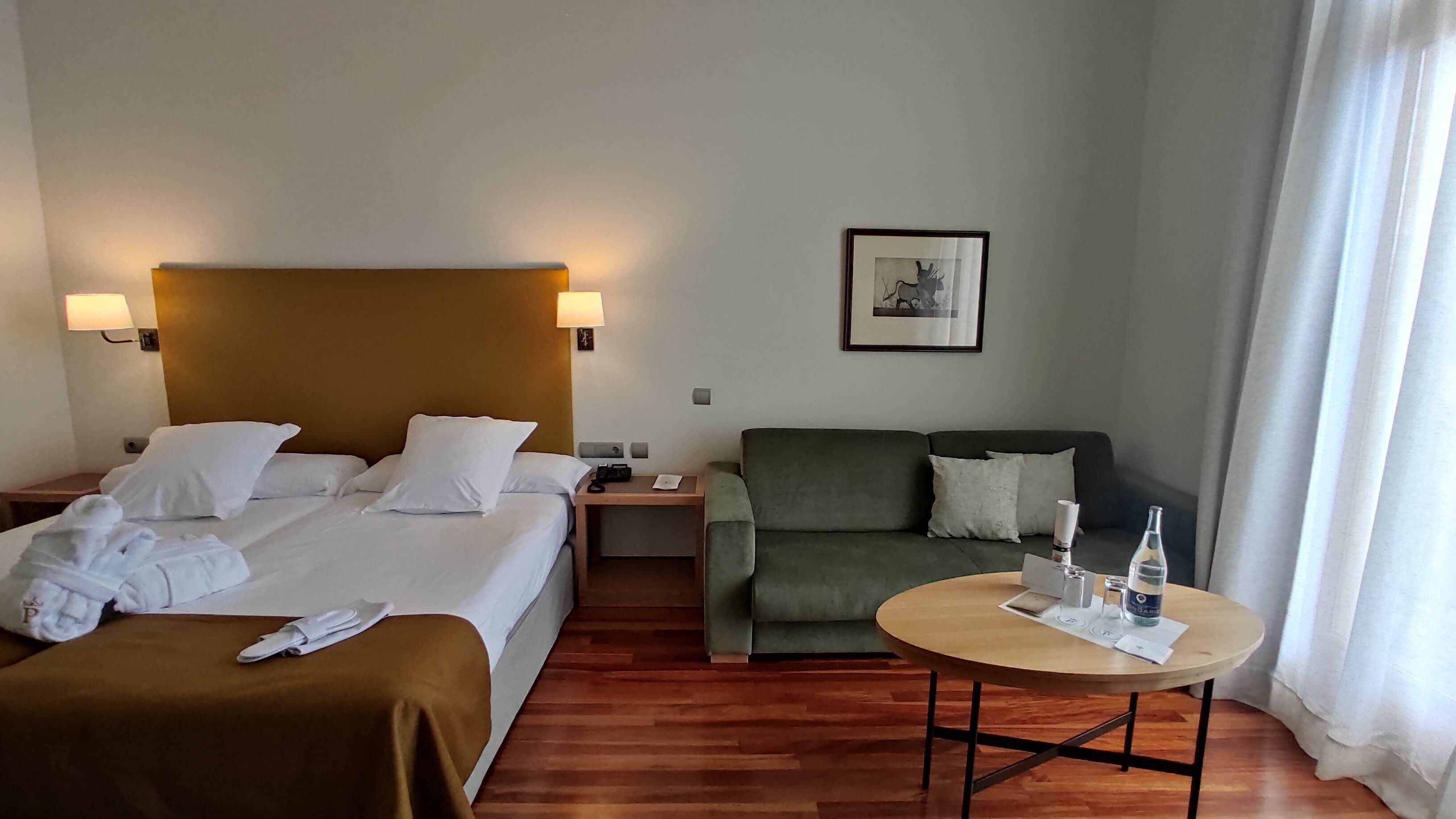 Une chambre d’hôtel avec deux lits, un canapé vert, une table ronde en bois et un tableau encadré au mur.