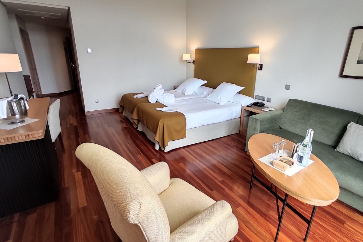 A hotel room with a bed, a green sofa, a small table with a bottle and glasses, a chair, and a desk with a lamp.