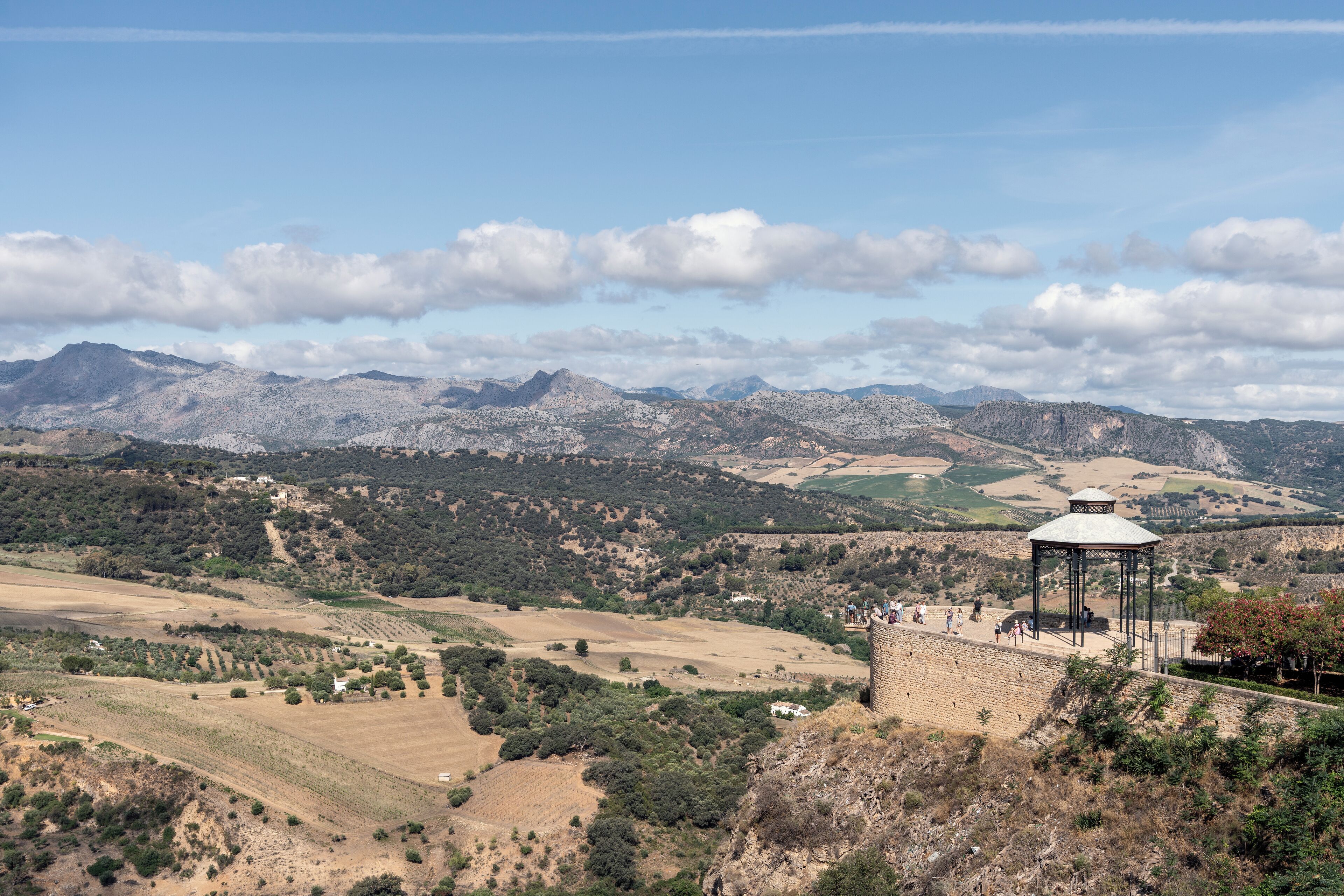 Weitläufige Aussicht auf eine gebirgige Landschaft mit einer Steinstruktur im Vordergrund, vermutlich eine Aussichtsplattform, umgeben von Feldern und spärlicher Vegetation.