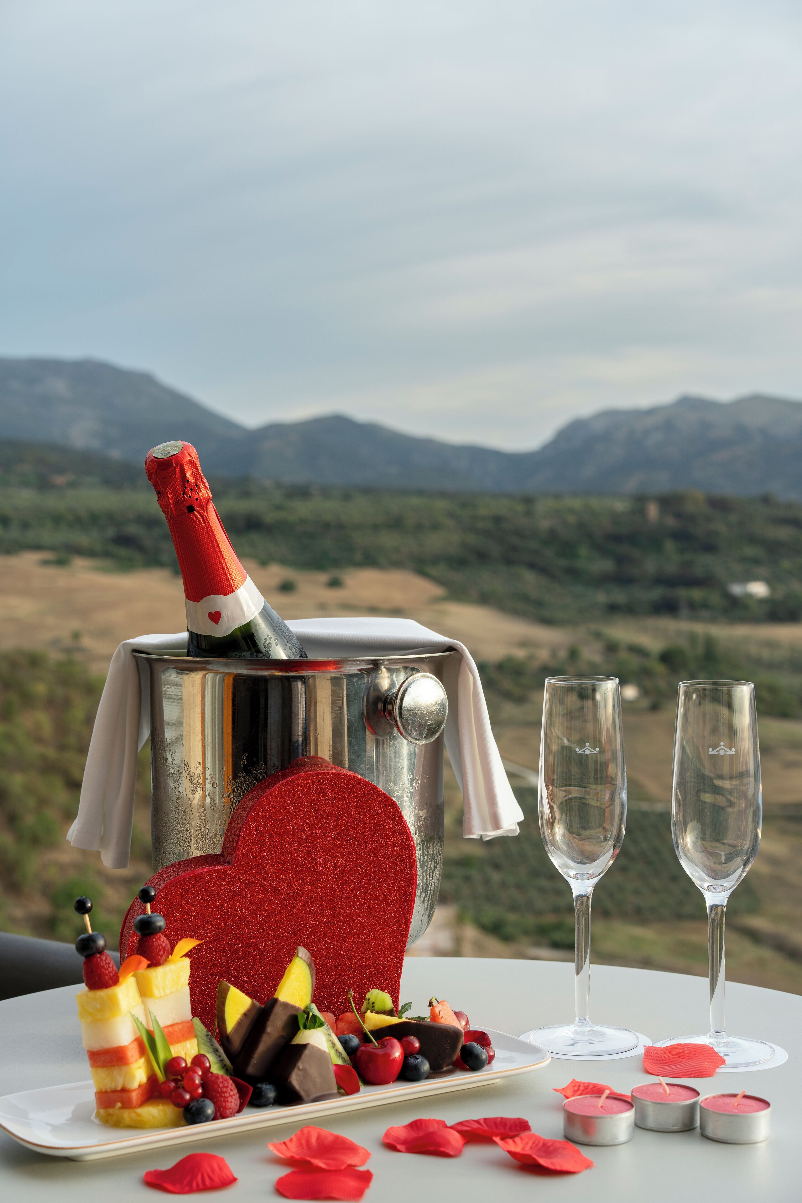 A table with a champagne bottle, fruit, and glasses, set against a mountainous backdrop.