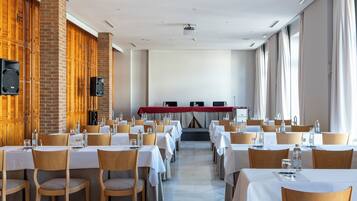 Une salle de conférence avec des rangées de tables et de chaises, une scène dotée d’un fond rouge et un mur de briques avec des casiers en bois.