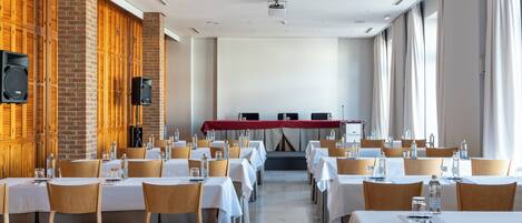 A conference room with rows of tables and chairs, a stage with a red backdrop, and a brick wall with wooden lockers.