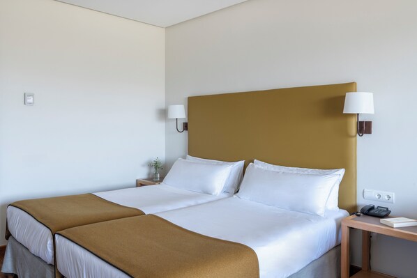 A double bed with white linens and a brown throw blanket, a bedside table with a lamp, and a wall socket.