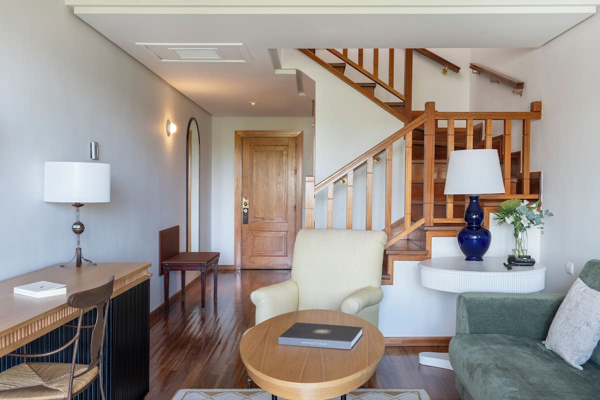 A spacious living room with a wooden staircase, a round coffee table, a sofa, and a dining area with a table and chairs.