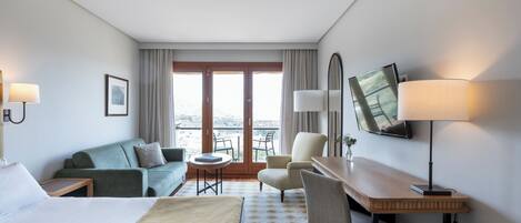 A hotel room with a bed, two armchairs, a desk, a TV, and a balcony with a view.