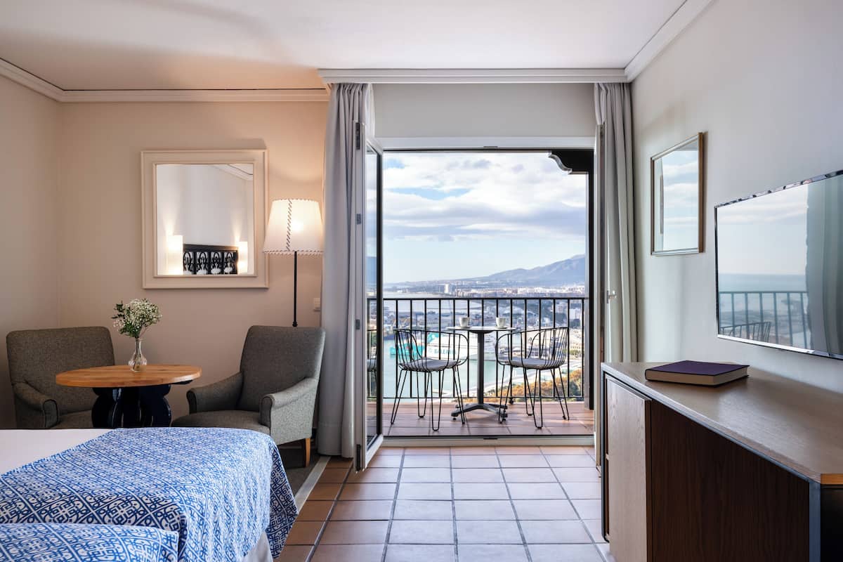 A hotel room with a bed, a desk, a chair, a small table with a vase, a balcony with a view of the sea and mountains, and a TV on the wall.