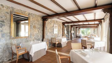 Un restaurante con paredes de piedra, vigas de madera y suelo de baldosas. Las mesas están dispuestas con manteles blancos y sillas.