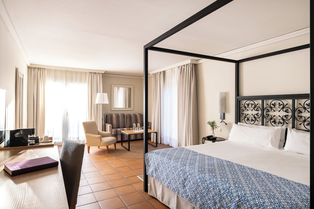 A hotel room with a four-poster bed, a sofa, a desk with a lamp, and a television.