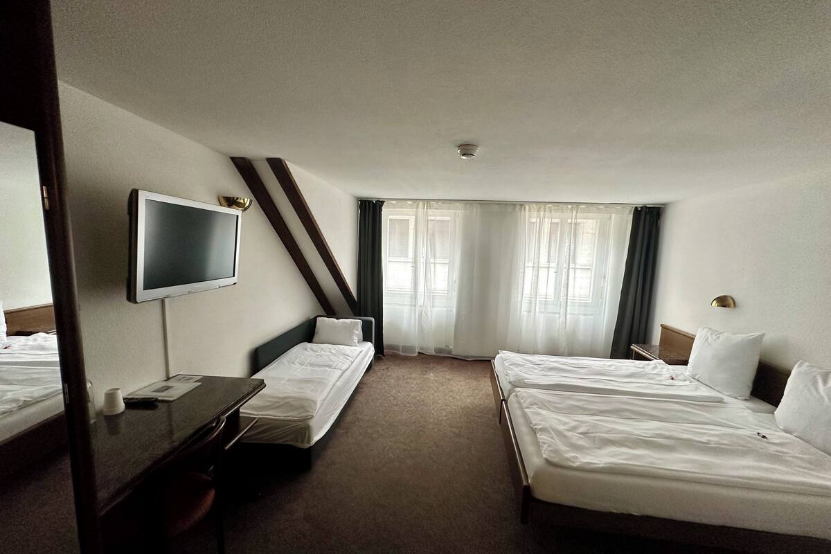 A hotel room with two single beds, a desk with a chair, a flat-screen TV, and a large window with curtains.