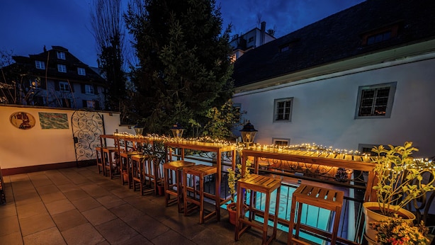 A poolside area with wooden tables and chairs, surrounded by potted plants and string lights. - BASLERTOR Summer Pool Hotel (Lucerne)