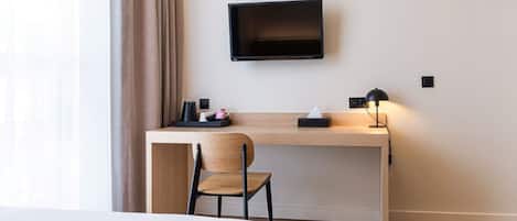 Habitación de hotel con escritorio de madera, una silla, televisor montado en la pared, mesita de noche con lámpara y cama.