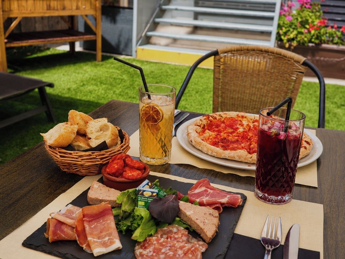 Un montaje para comer al aire libre con pizza, una bebida, pan y varias carnes sobre una mesa.