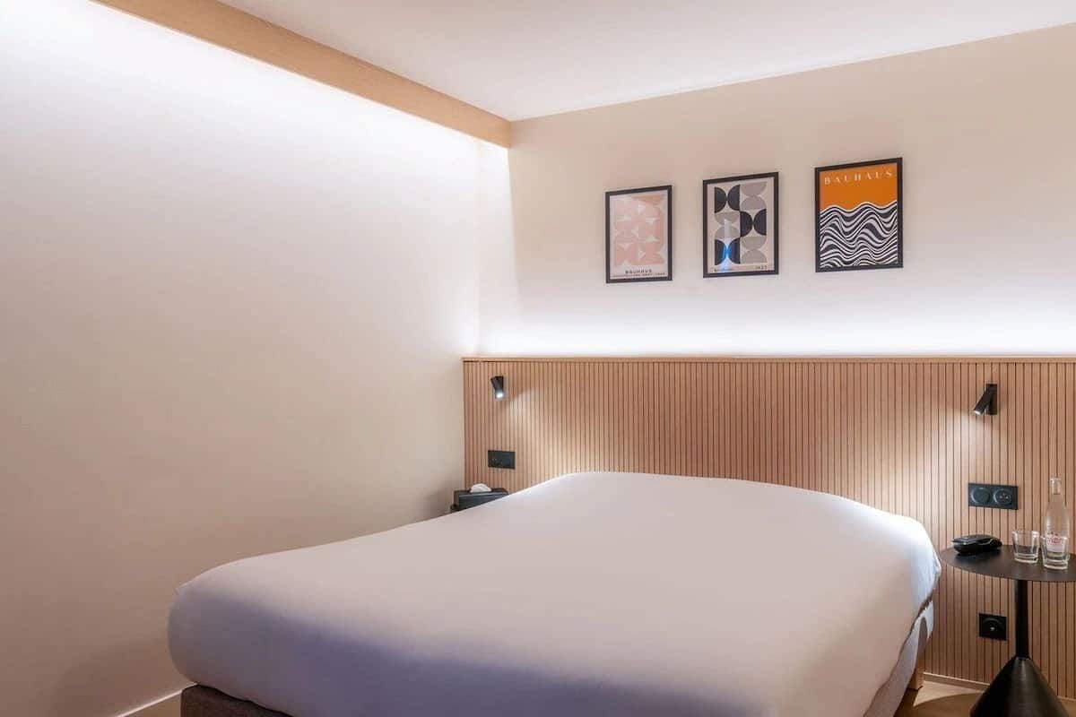 Een hotelkamer met een bed, een houten hoofdeinde, een nachtkastje en drie ingelijste kunstwerken aan de muur.