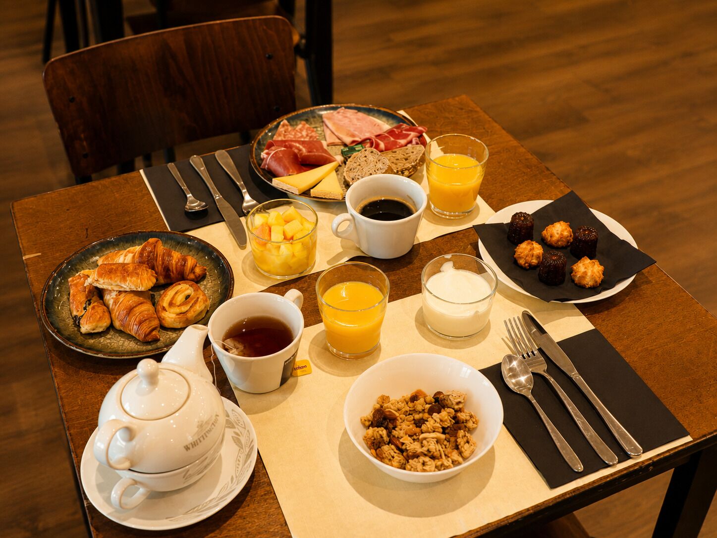 Une table de petit-déjeuner avec des croissants, un bol de céréales, une théière, une tasse de café, un verre de jus d’orange et une assiette de charcuterie.