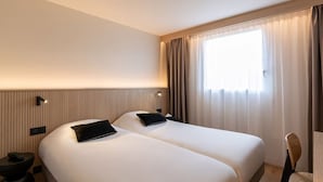 A hotel room with two beds, a desk, a chair, and a large window with curtains. - Grand Hôtel de la Seine (Rouen)