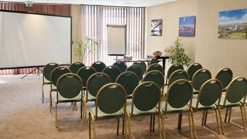Un salón de conferencias con filas de sillas, una pantalla de proyector, una mesa con una planta y cuadros en la pared.