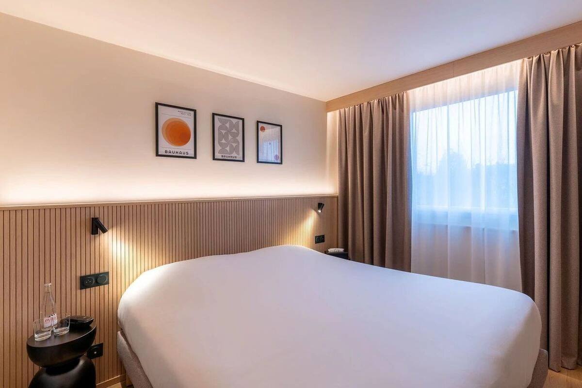 Een hotelkamer met een groot bed, een nachtkastje met een lamp en een raam met gordijnen.