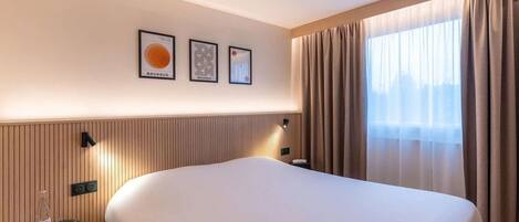 A hotel room with a large bed, a bedside table with a lamp, and a window with curtains.