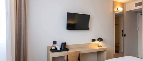 Habitación de hotel con cama, escritorio con silla, televisor de pantalla plana montado en la pared, lámpara de noche y una mesita con cafetera y tazas.