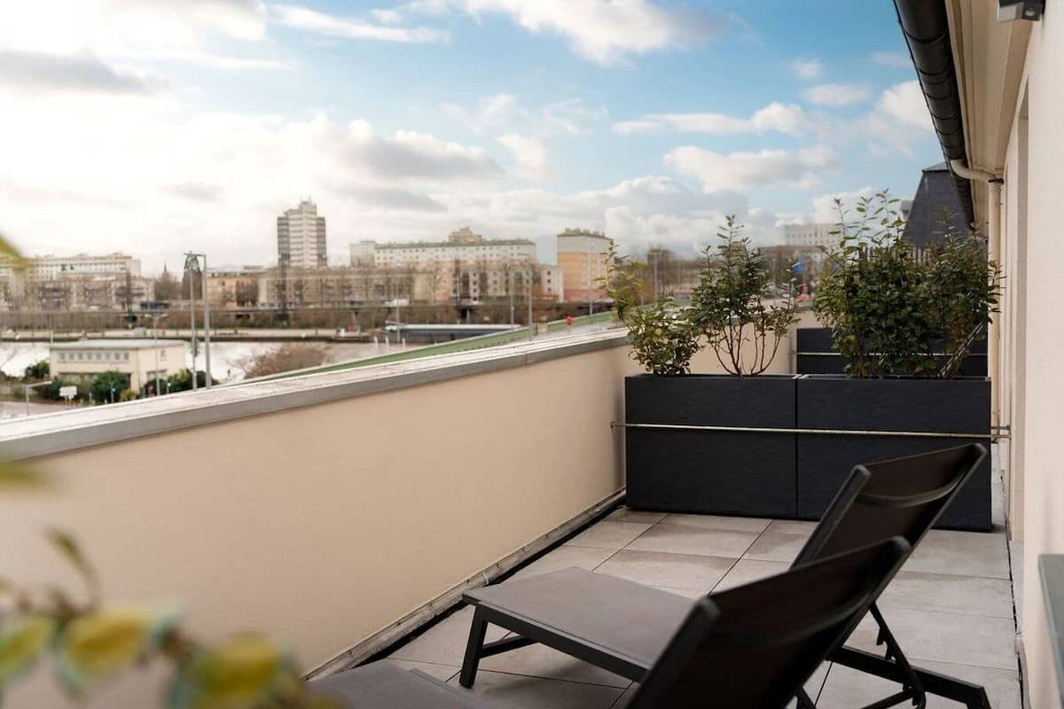 Een balkon met uitzicht op een stadsgezicht, met gebouwen en een brug. Er staan twee zwarte stoelen en een plantenbak met planten.