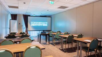 Un salón de conferencias con mesas y sillas, una pantalla de proyector que muestra "GRAND HOTEL DE SEINE", y una pizarra.