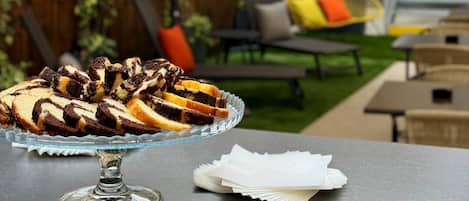 A dessert platter on a table with a glass stand, a white napkin, and an outdoor seating area with a yellow chair and a white umbrella.