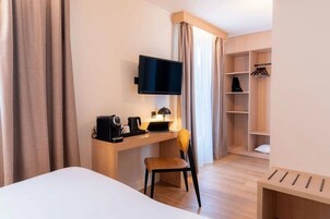 Habitación de hotel con cama, escritorio con cafetera, silla, televisor montado en la pared y armario con espacio para colgar ropa.
