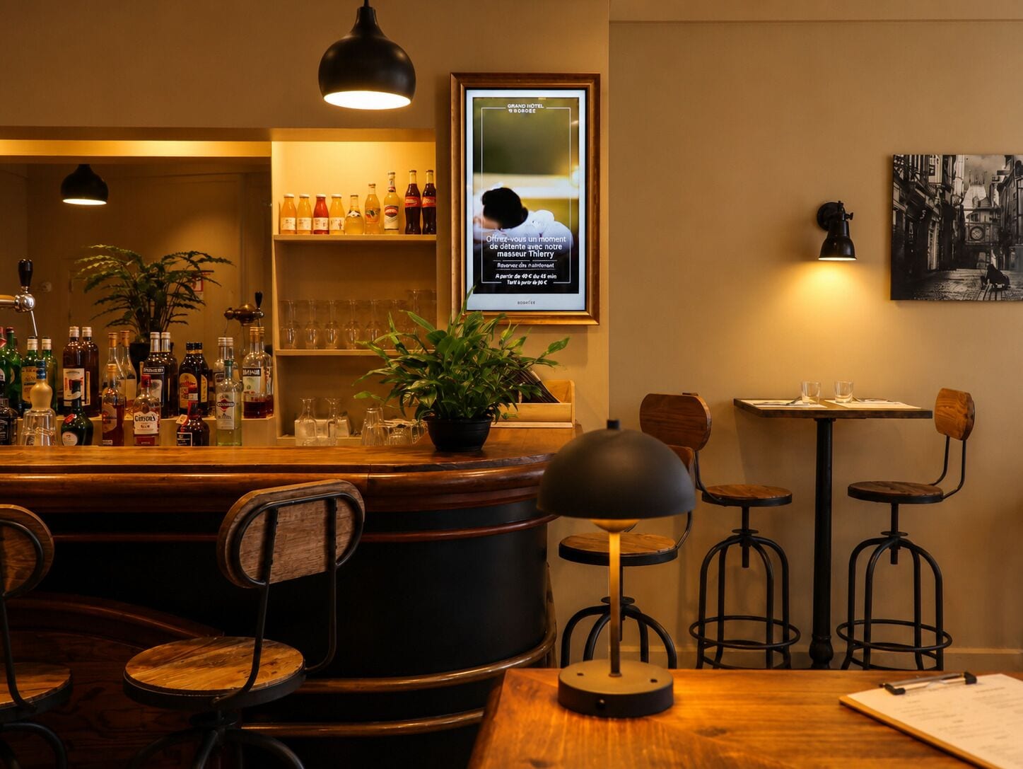 A bar area with a variety of bottles, glasses, and a potted plant. There is a framed poster on the wall and a table with chairs.