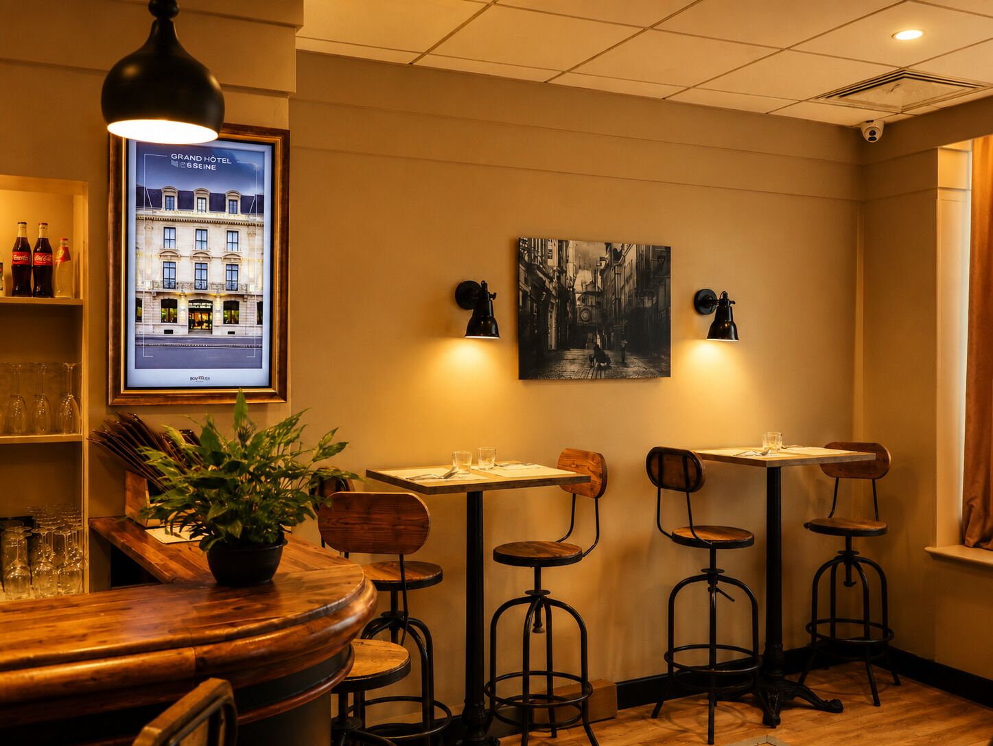 Interior de un restaurante con barra, mesas y sillas de madera, una planta en maceta y obras de arte en la pared.