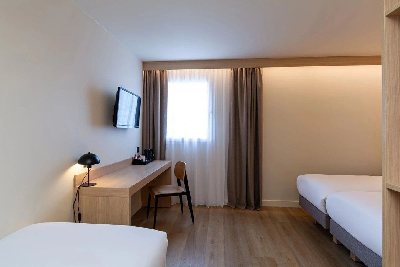 Una habitación de hotel con dos camas individuales, un escritorio con silla, un televisor montado en la pared y una ventana con cortinas.