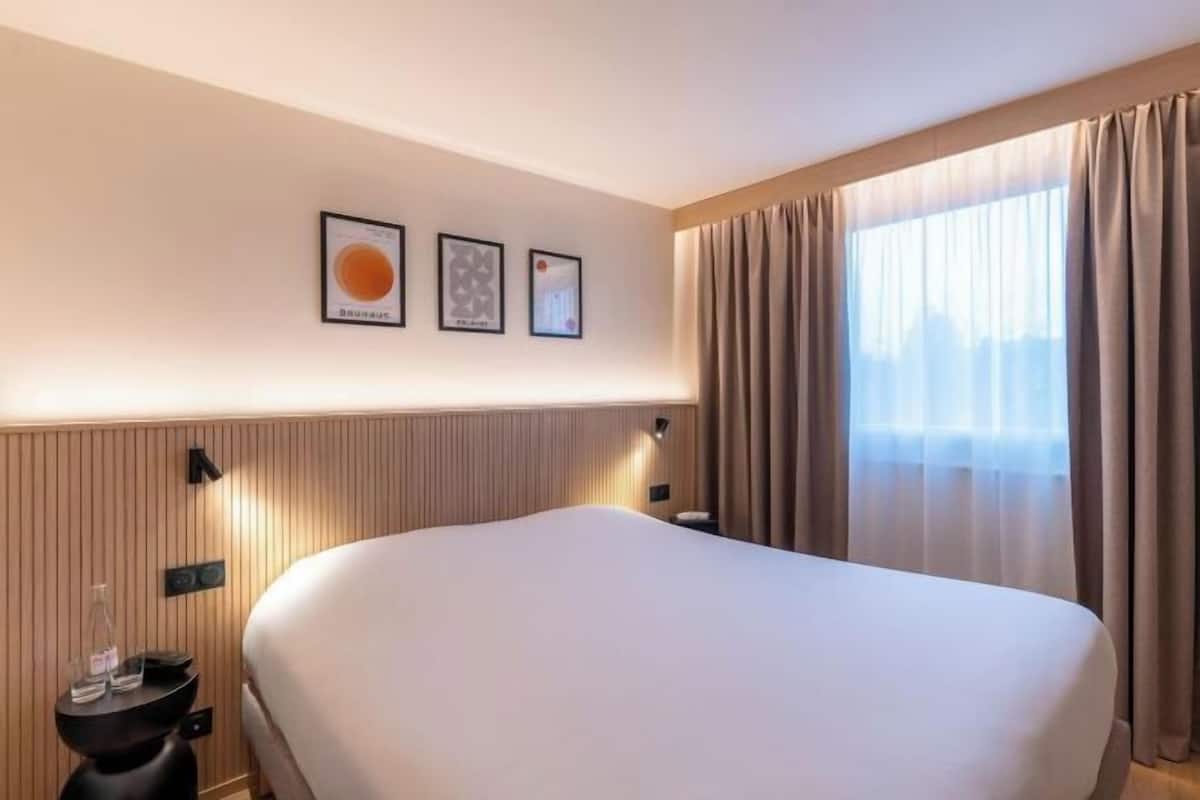 Een hotelkamer met een groot bed, een nachtkastje met een fles en glazen, een wandlamp en een raam met gordijnen.