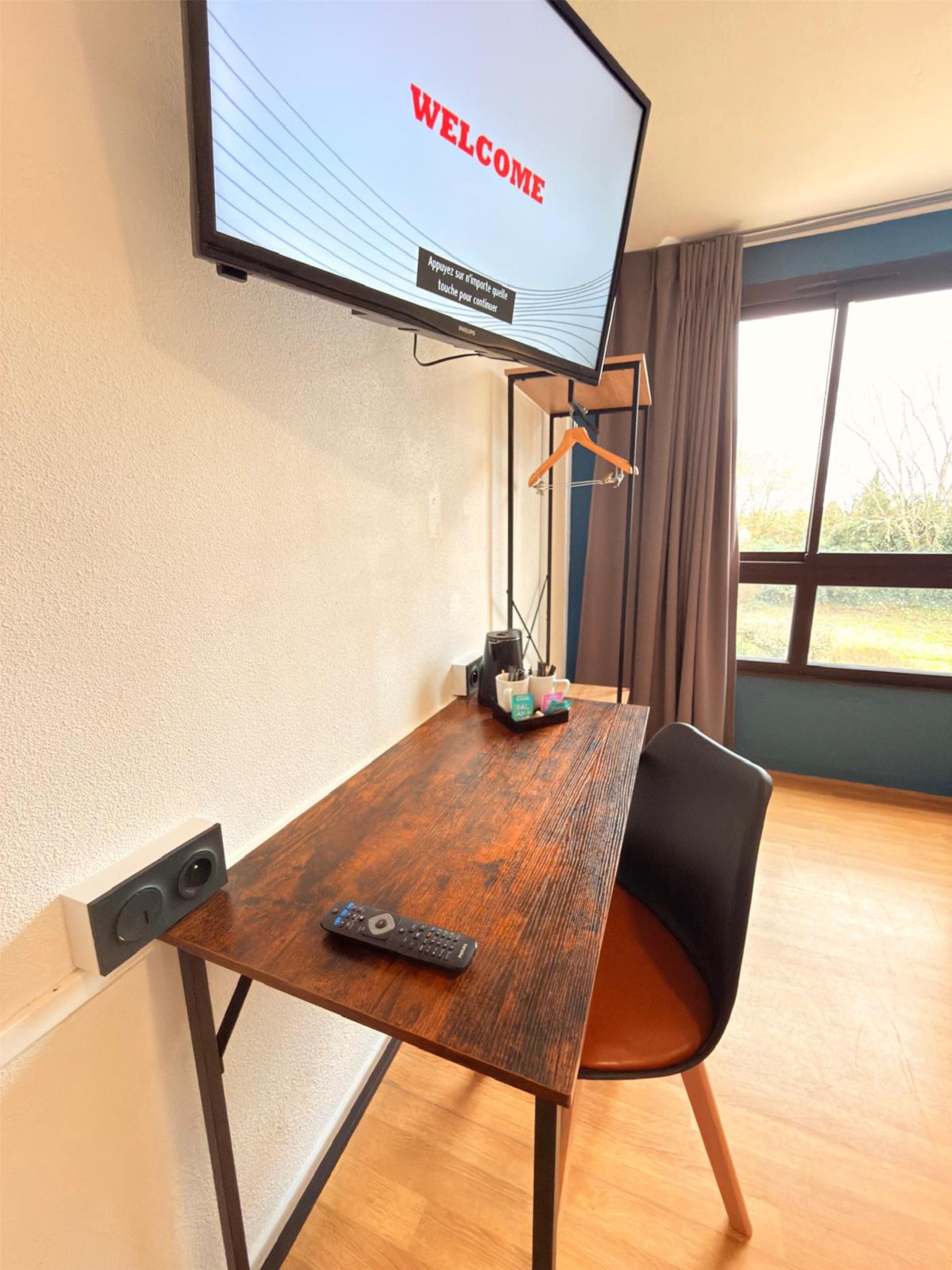 A room with a wooden table, a black chair, a TV displaying "WELCOME" on the screen, and a window with curtains.