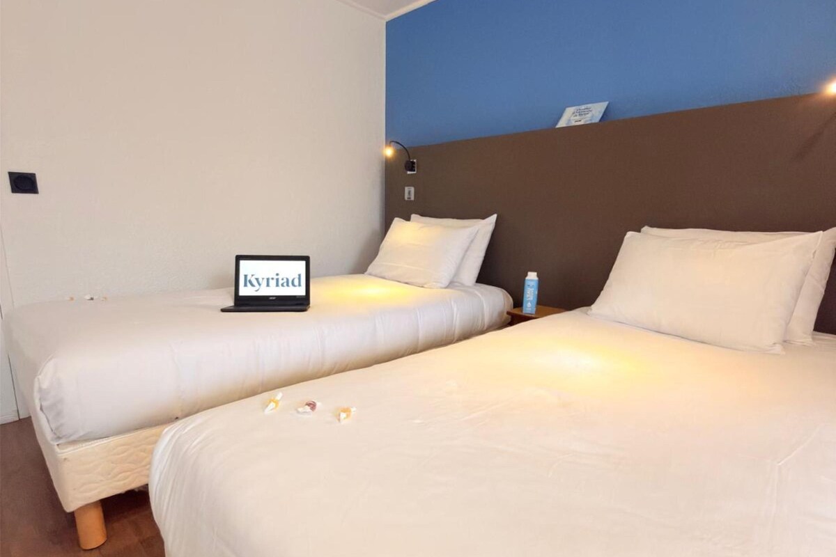 A hotel room with two beds, a blue accent wall, and a sign on the left bed reading "Kyriad."