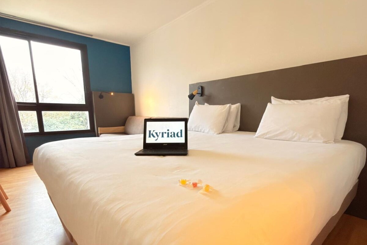 A hotel room with a large bed, a laptop displaying 'Kyriad', and a view through a large window.