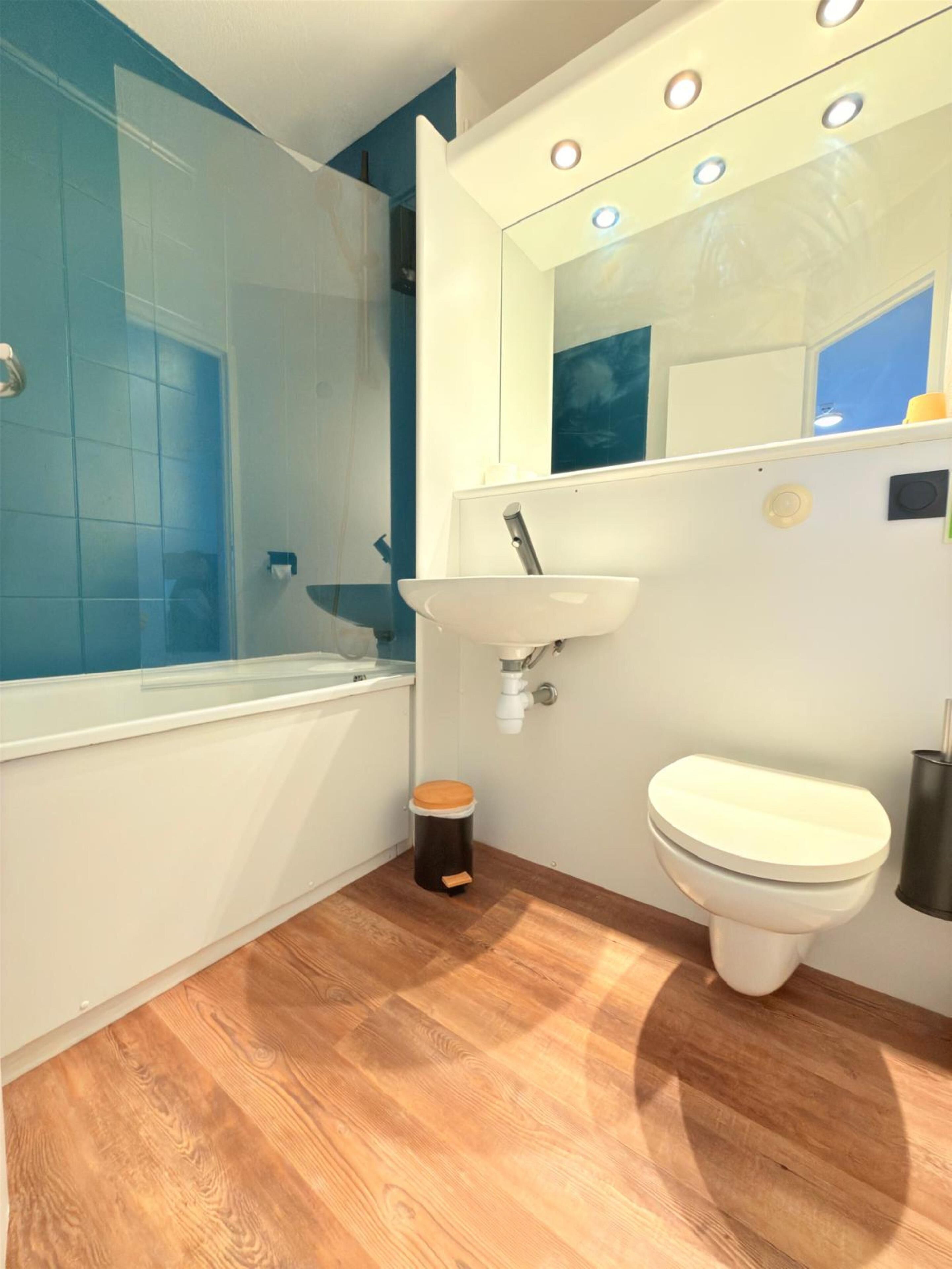 A modern bathroom with a wooden floor, a white sink, a toilet, and a shower area with blue tiles.